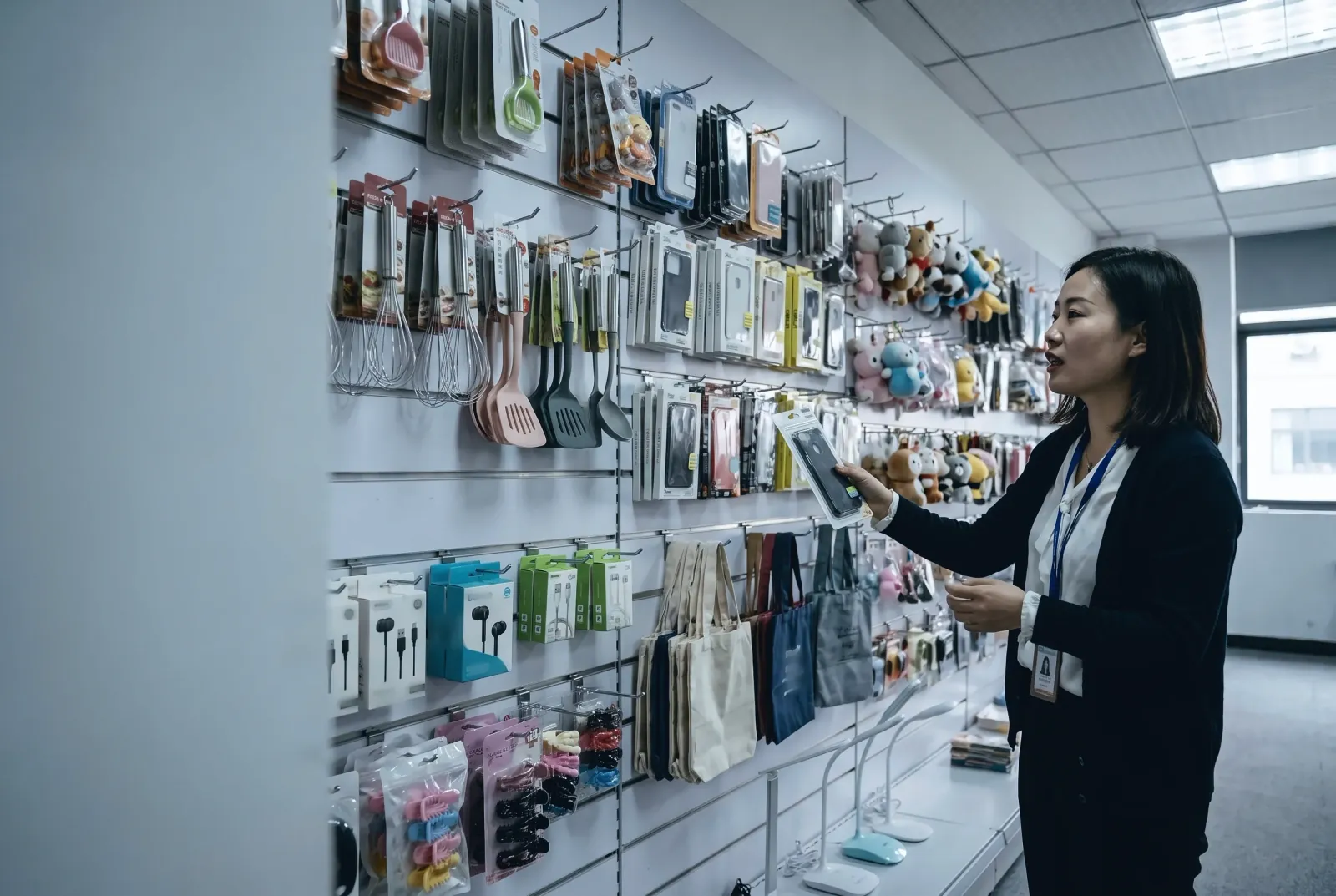 Une acheteuse examine un article dans le showroom d'un grossiste chinois, murs couverts d'ustensiles de cuisine, d'écouteurs et de peluches